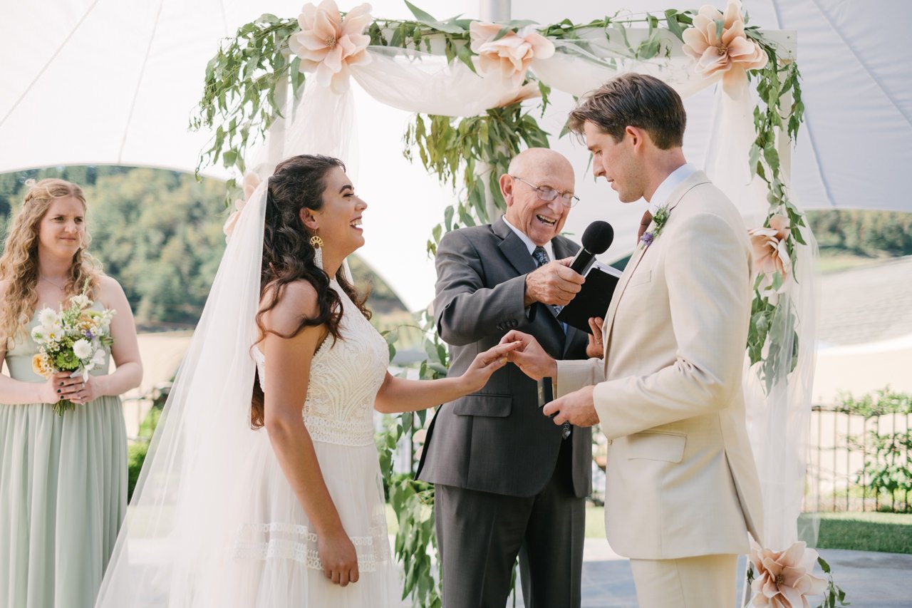  Bride laughs as groom in tan suit slips ring on her finger 