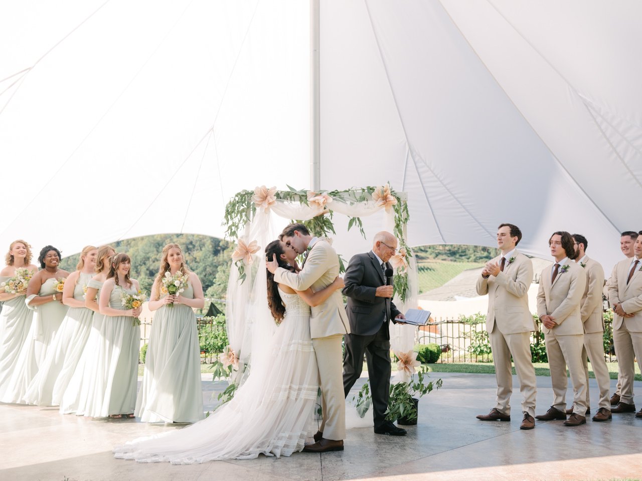  Bride and groom kiss under pink floral backdrop while wedding party in tan and green cheers under sun tent 