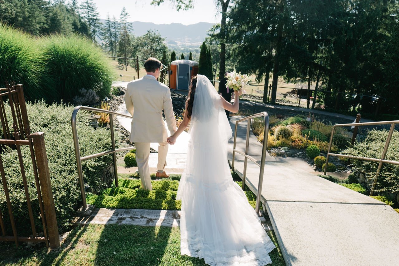  Bride and groom walk down grass steps backlit by sunlight  and valley 