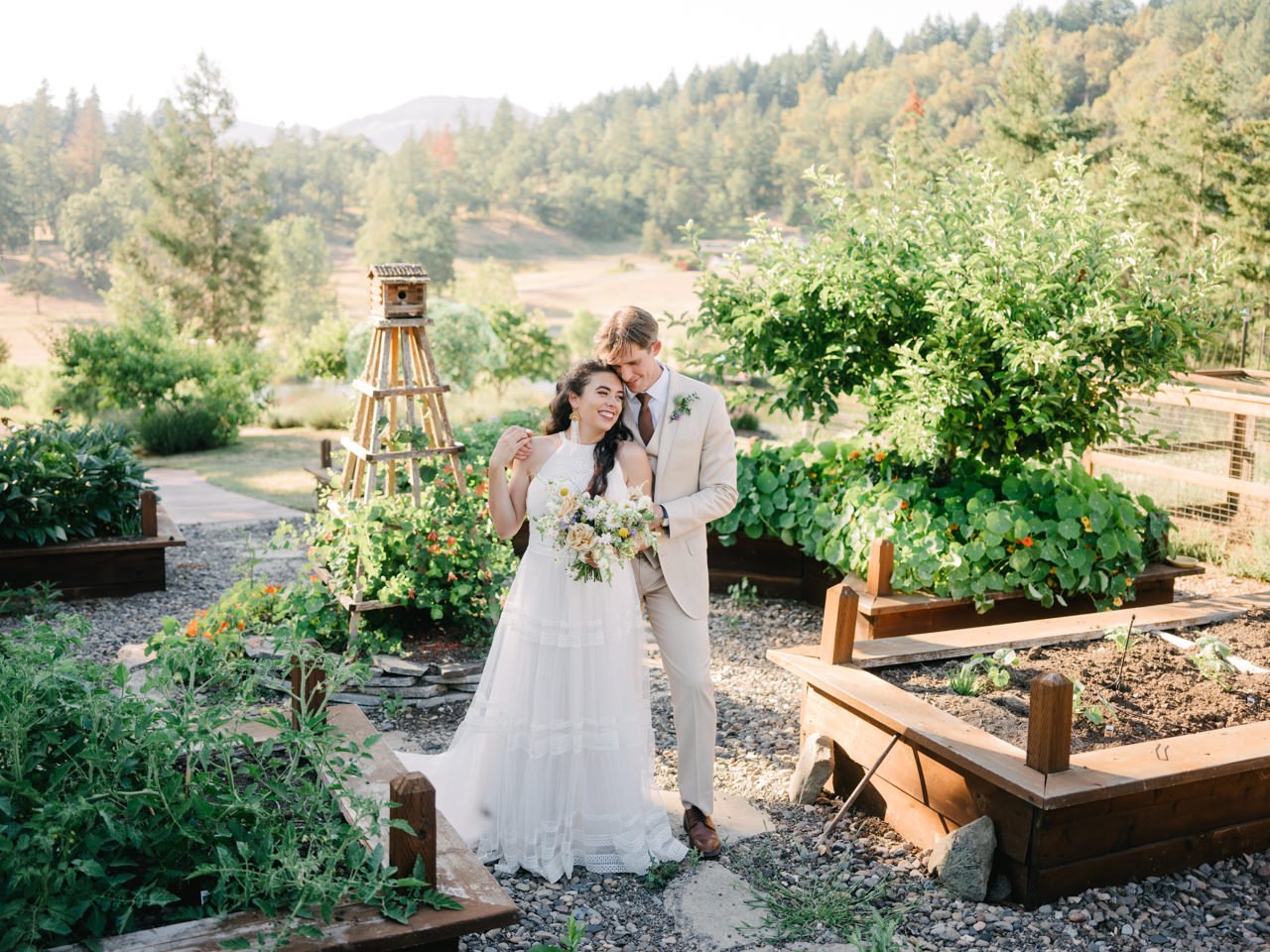  Bride and groom standing together in garden of raised beds and birdhouse with southern Oregon valley in background 