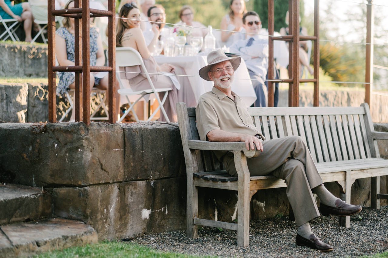  Father of bride wearing tan green brim hat sits on bench and laughs 