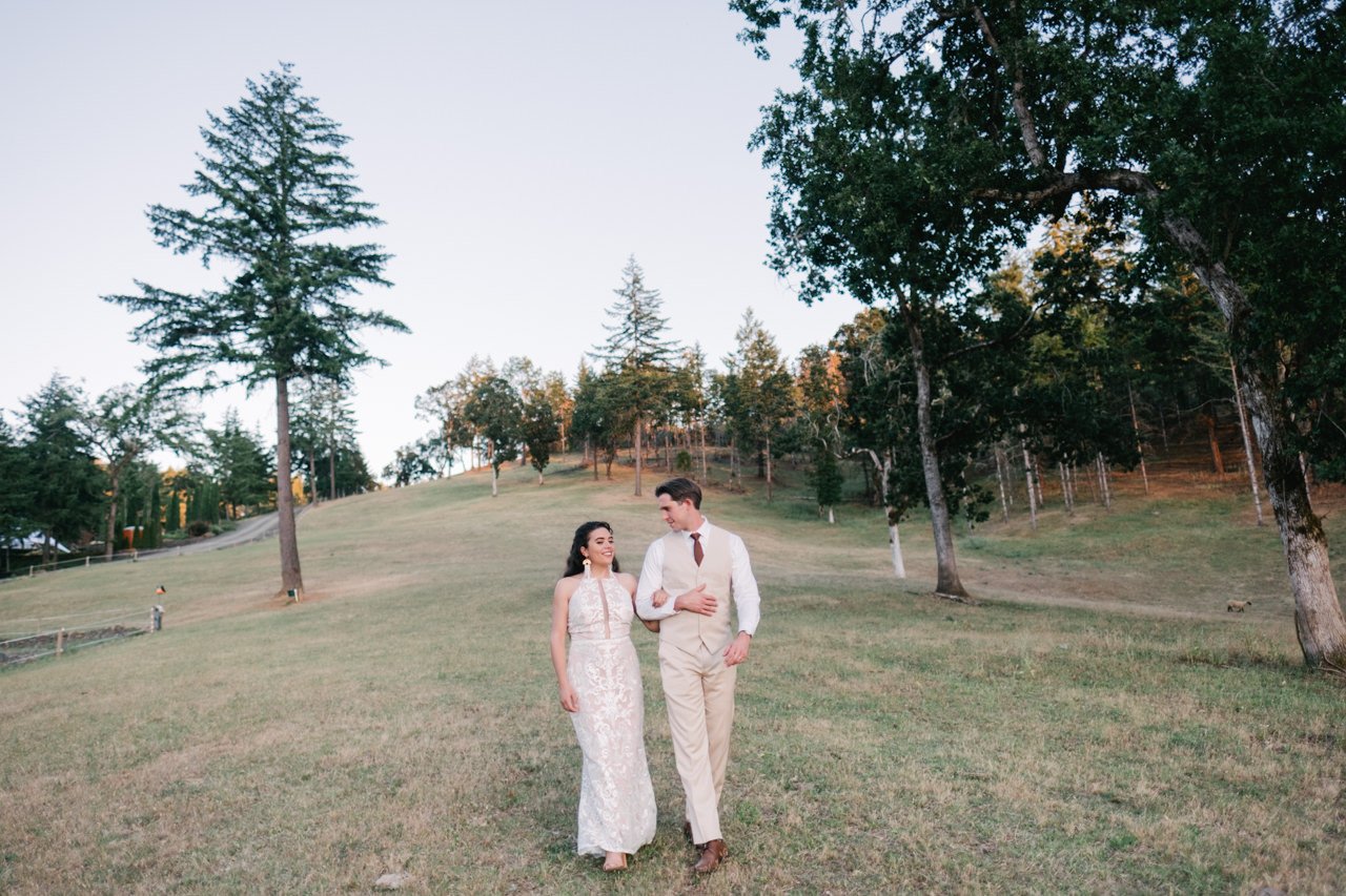  Bride and groom walk down farm pasture slope evening 