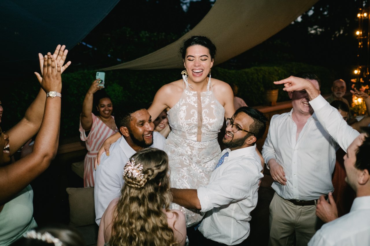  Bride held up laughing on shoulders during dance while guests clap 
