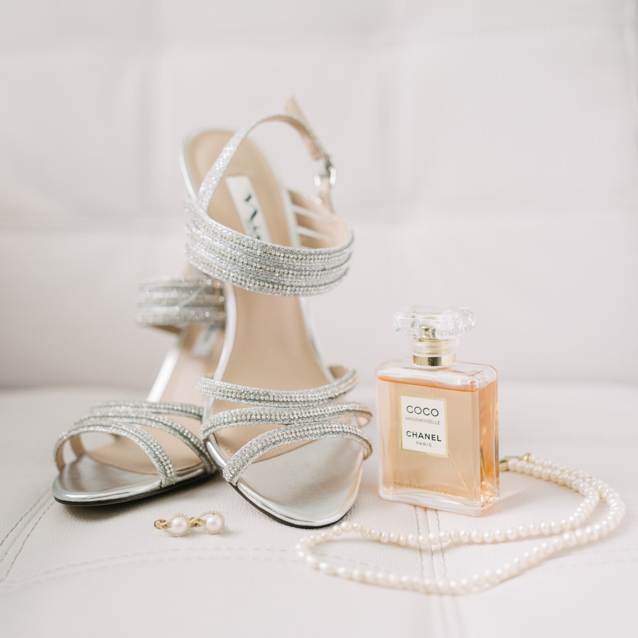  Detail photo of bride shoes, necklace, coco Chanel perfume and earrings on white chair 