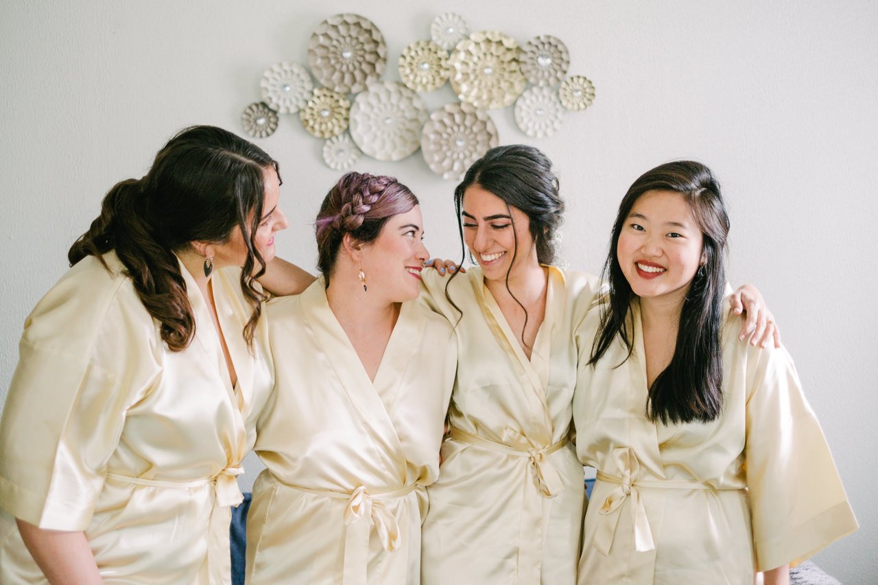  Bridesmaids in gold bathrobes smile together in front of living room gold wall piece 