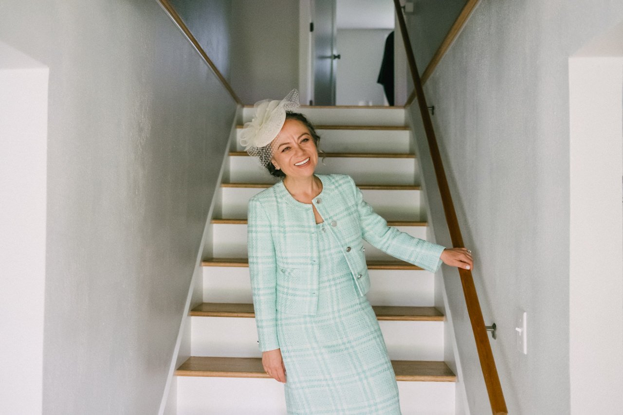  Mother of bride in teal outfit and hairpiece smiles in doorway steps 