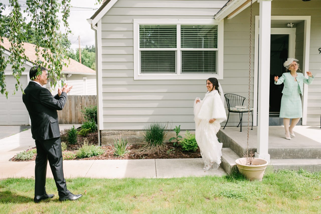  Bride in white wedding dress exits airbnb house while mother dances on steps and father claps 