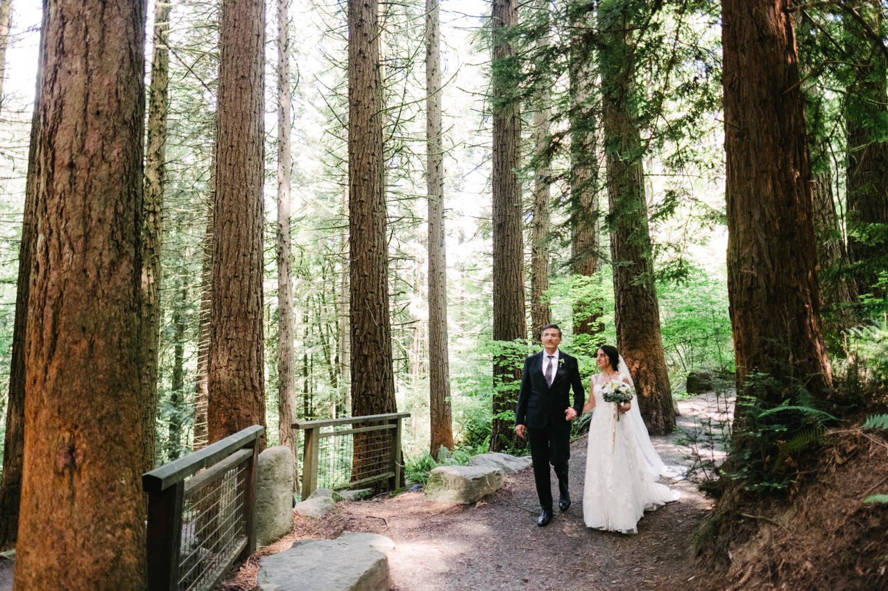 Bride in white dress walks with father down trail surrounded by redwood trees 