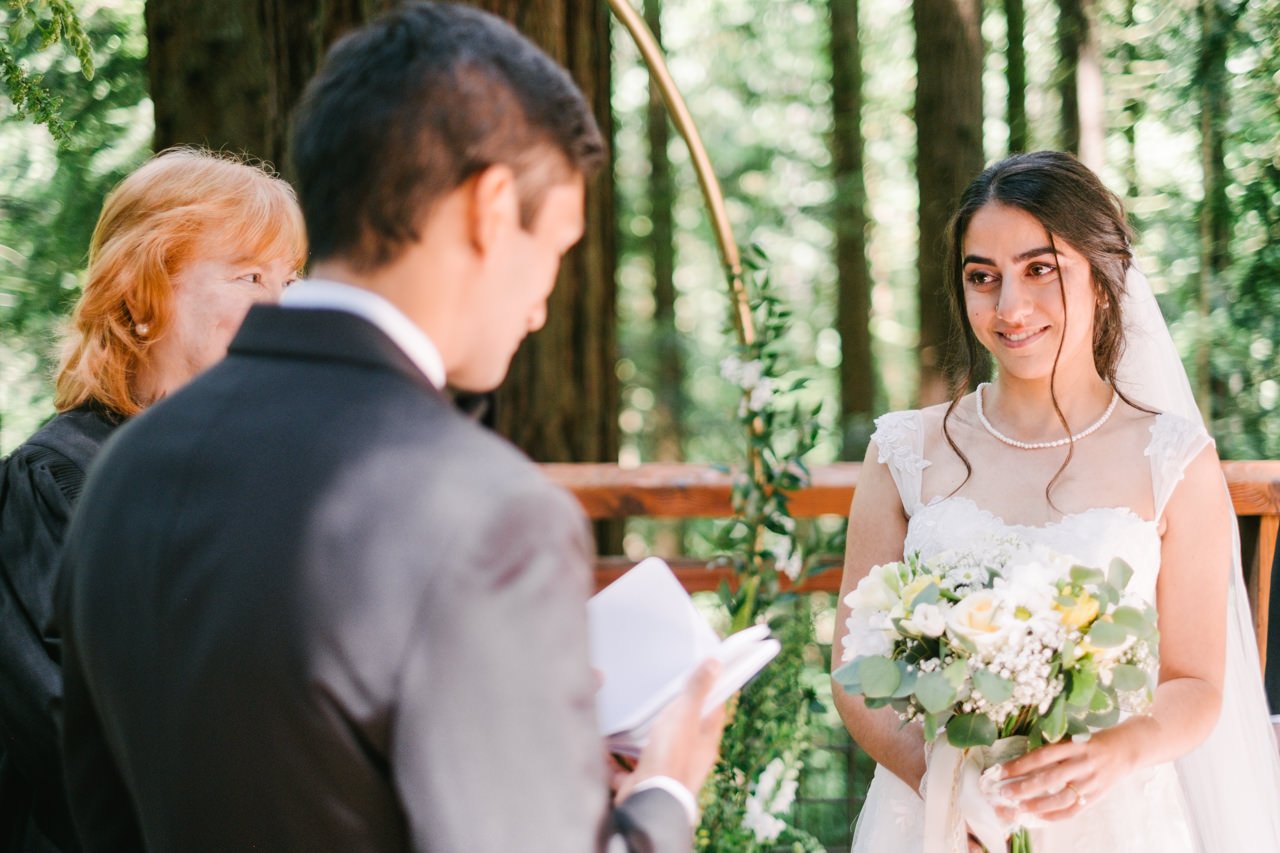  Bride with white and yellow bouquet smiles at groom while he reads vows 
