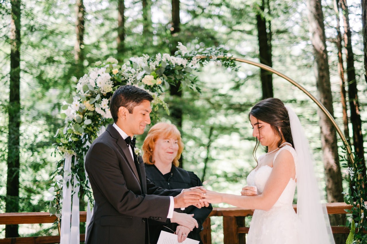  Groom places ring on brides finger with floral backdrop on redwood deck 