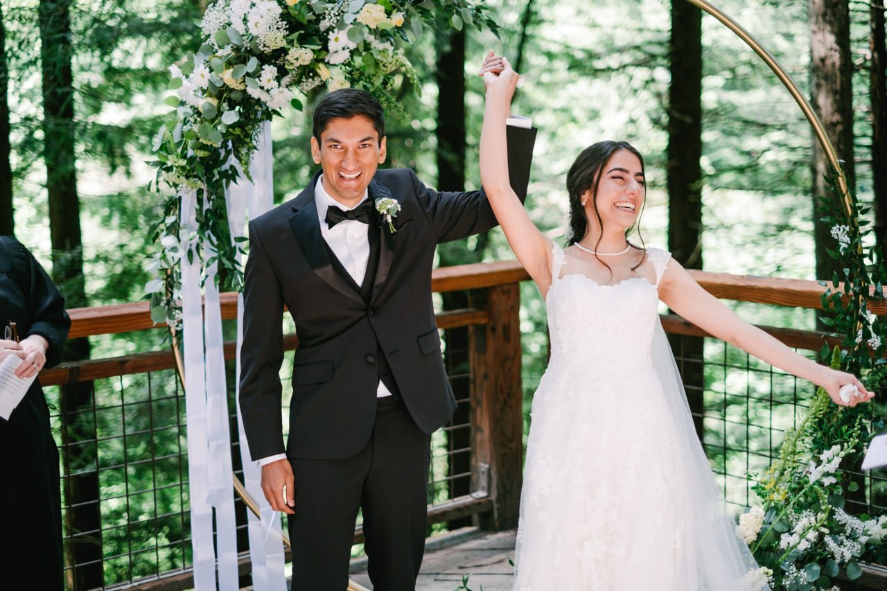  Bride and groom hold hands up and celebrate in front of circular floral backdrop on redwood deck 