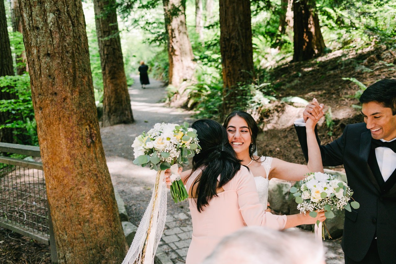  Bride hugs bridesmaid while holding grooms hand in forest trail 