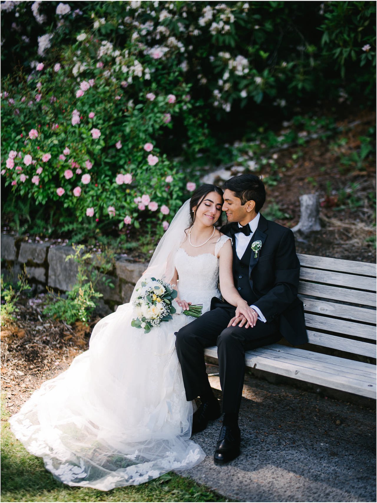 Bride and groom in tux sit on bench in rose test garden 