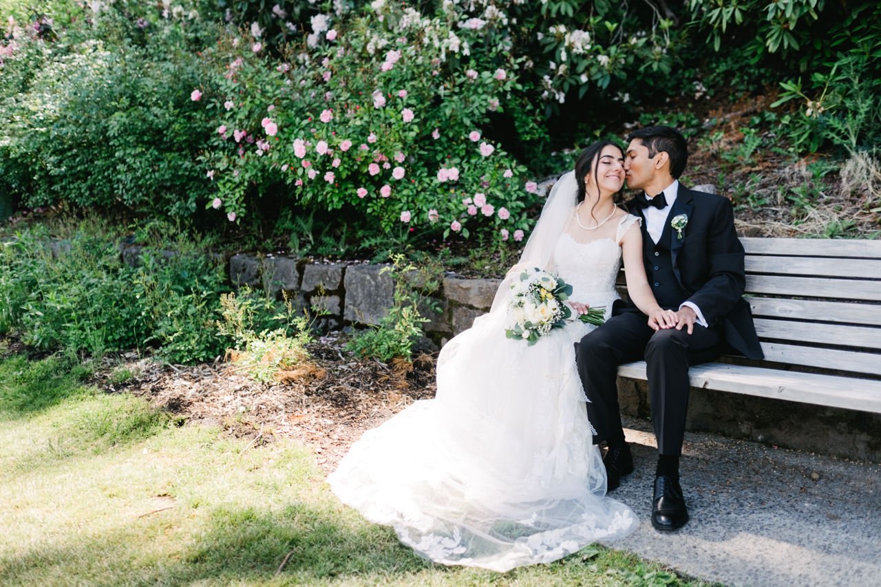  Groom kisses bride on bench in front of pink rose bush 