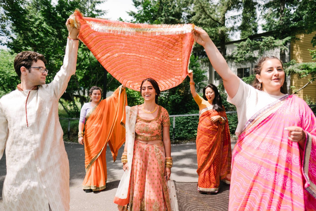  Bride smiles under orange covering during bridal Indian entrance 
