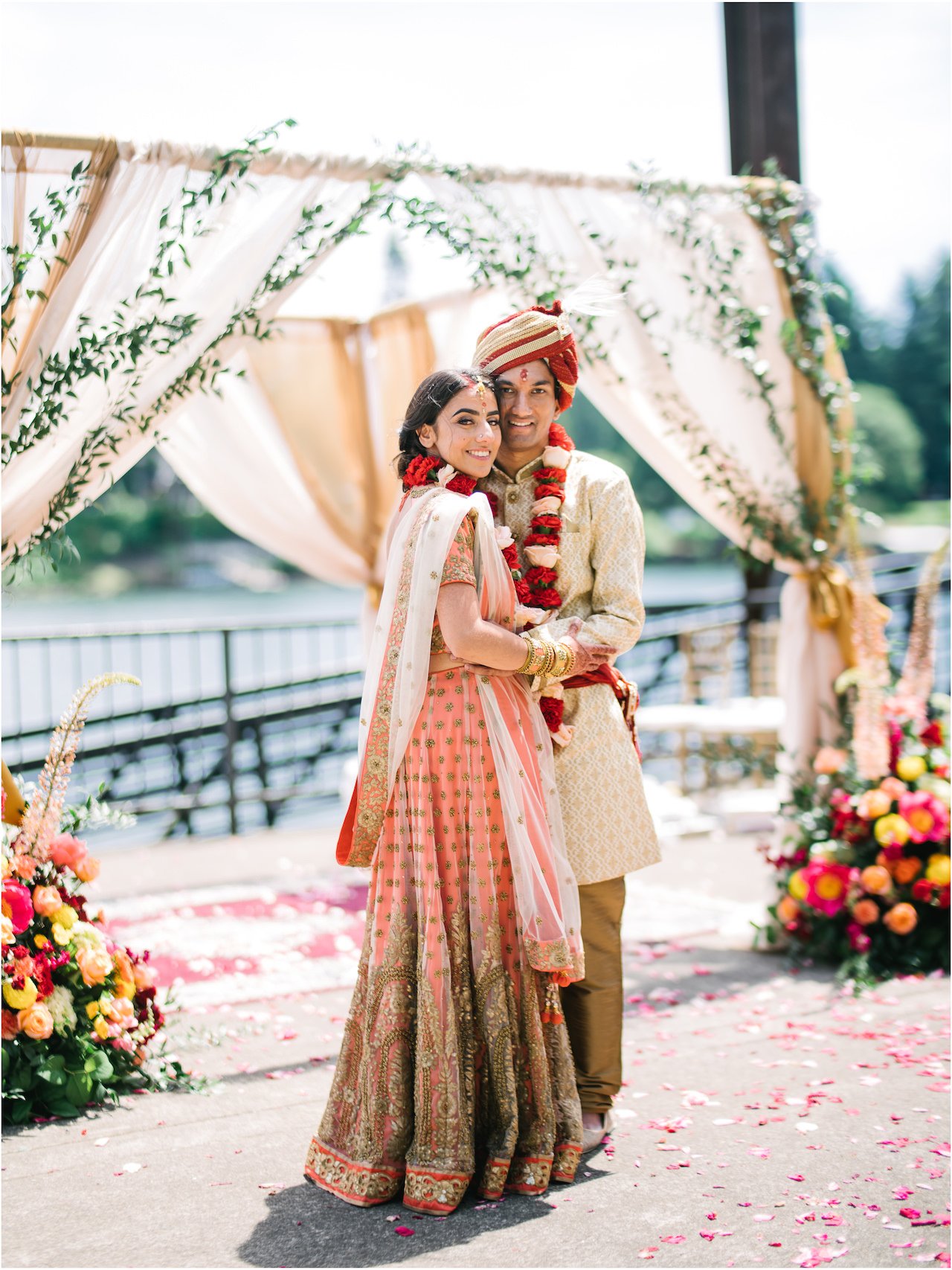  Bride and groom pose under floral mandap with red and white garland necklaces 