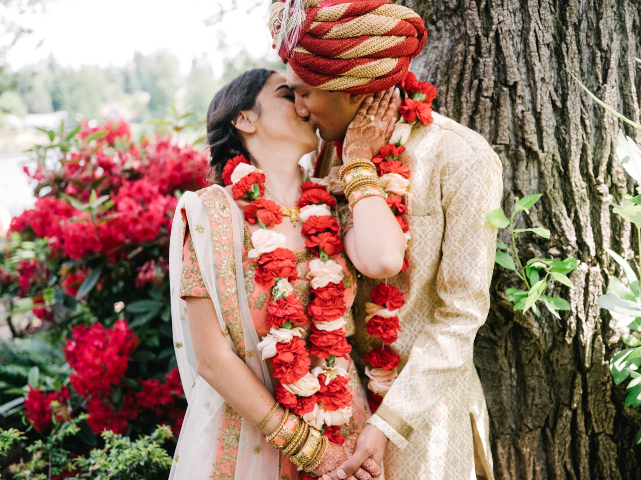  Indian couple shares kiss by tree with red and gold attire and red rhododendrons behind them 