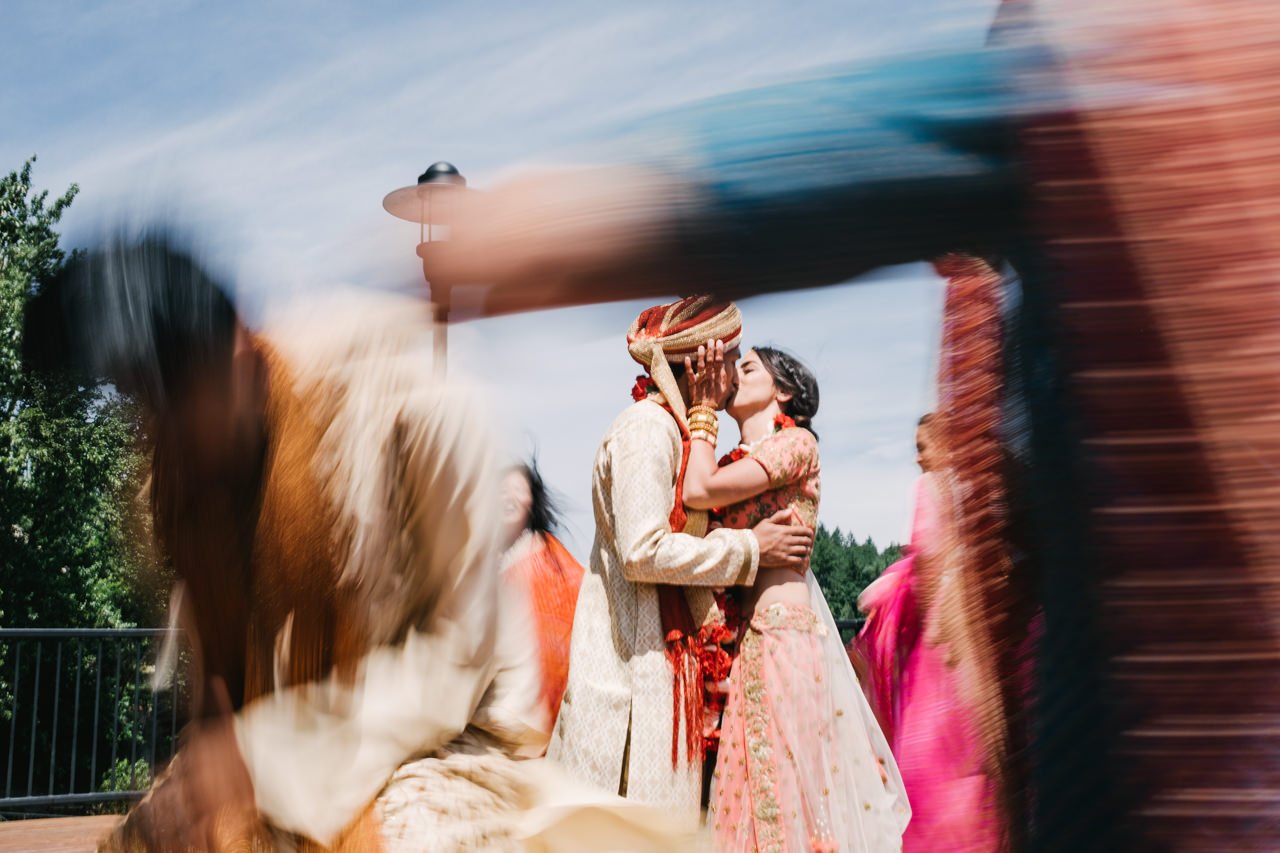  Indian couple in traditional attire share kiss while blurry wedding party dances around them 