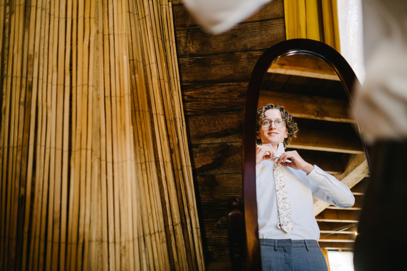  Groom smiling while adjusting floral tie in mirror next to bamboo frame 