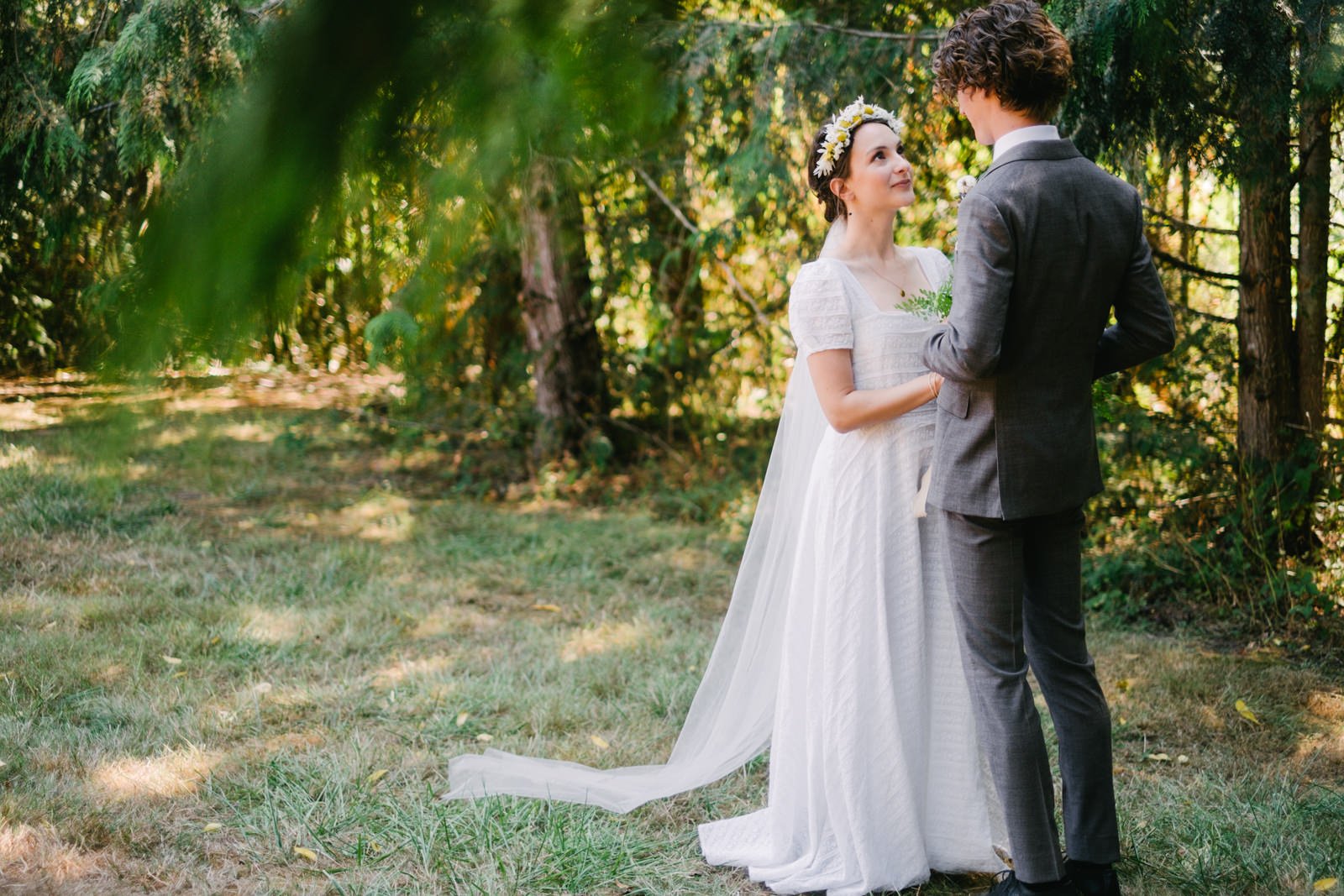  Bride with long veil looks into eyes of groom in grey suit surrounded by fir trees 