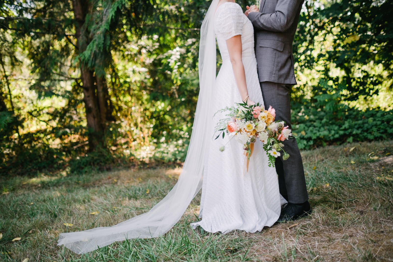  Bride and groom photo holding hands from shoulders down holding pink orange and fern bouquet with sunlight through the trees 