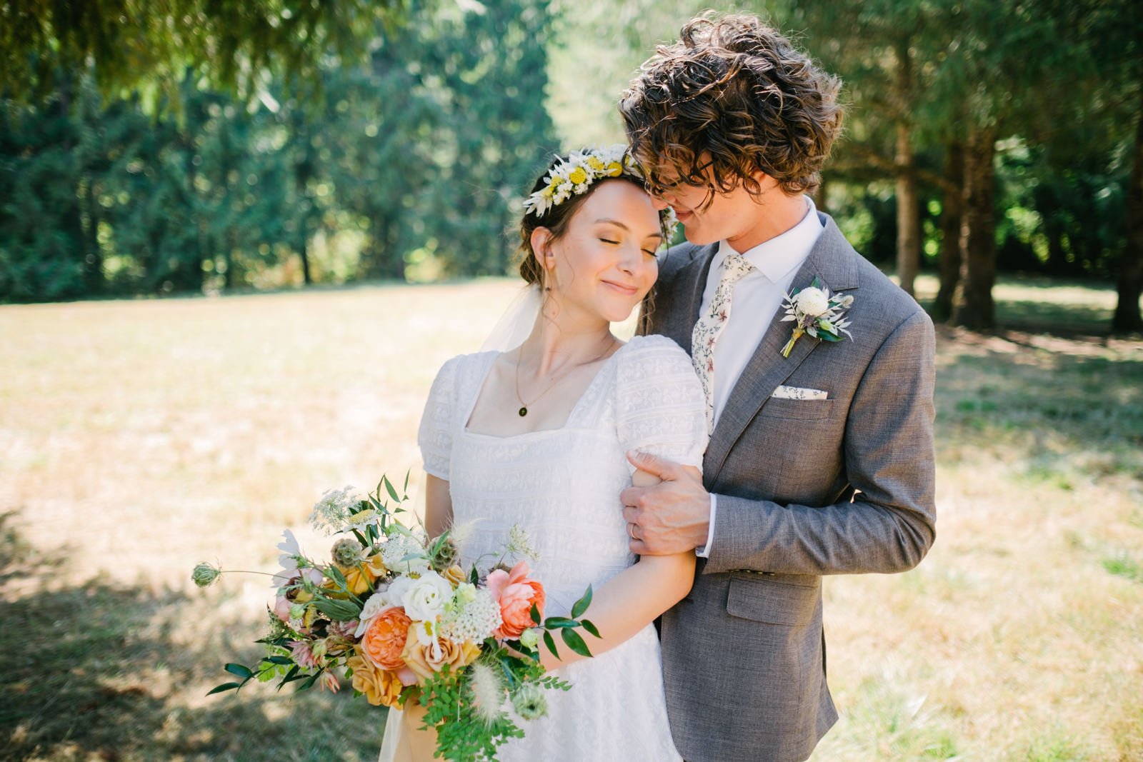  Bride with yellow daisy flower crown smiles while being embraced by groom in floral tie and grey suit in front of farm field 