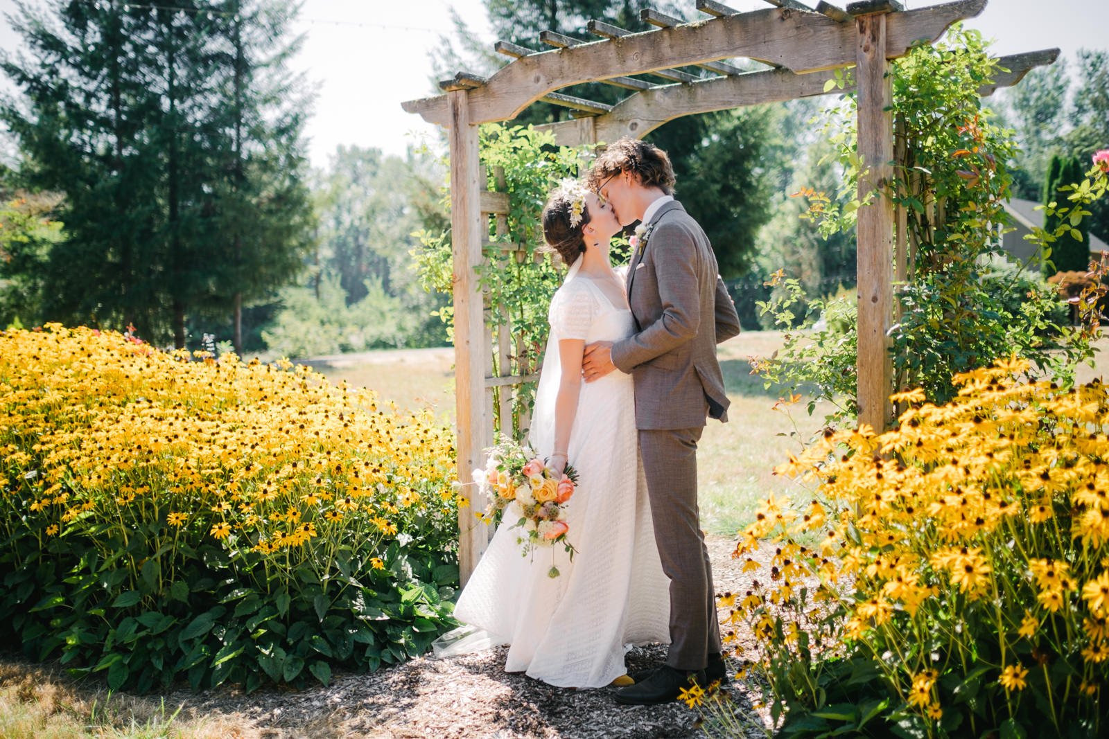  Bride and groom kiss under arbor surrounded by black eyed Susans holding colorful bouquet 