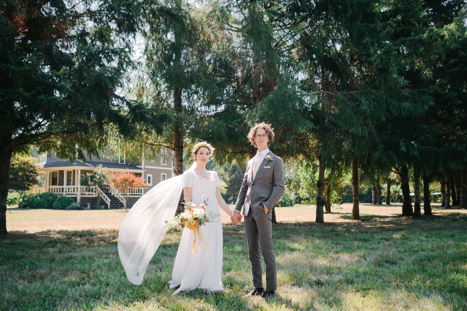  Bride and groom stand in field in front of trees and country house with veil blowing in breeze 