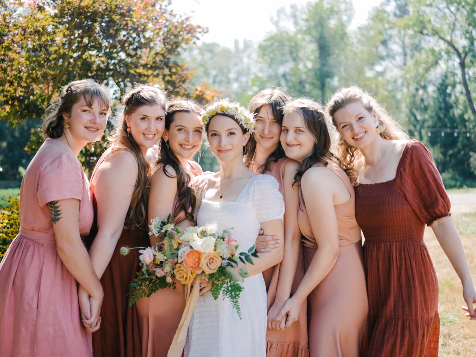  Bride with flower crown and bridesmaids in peach red and pink dresses snuggle together 