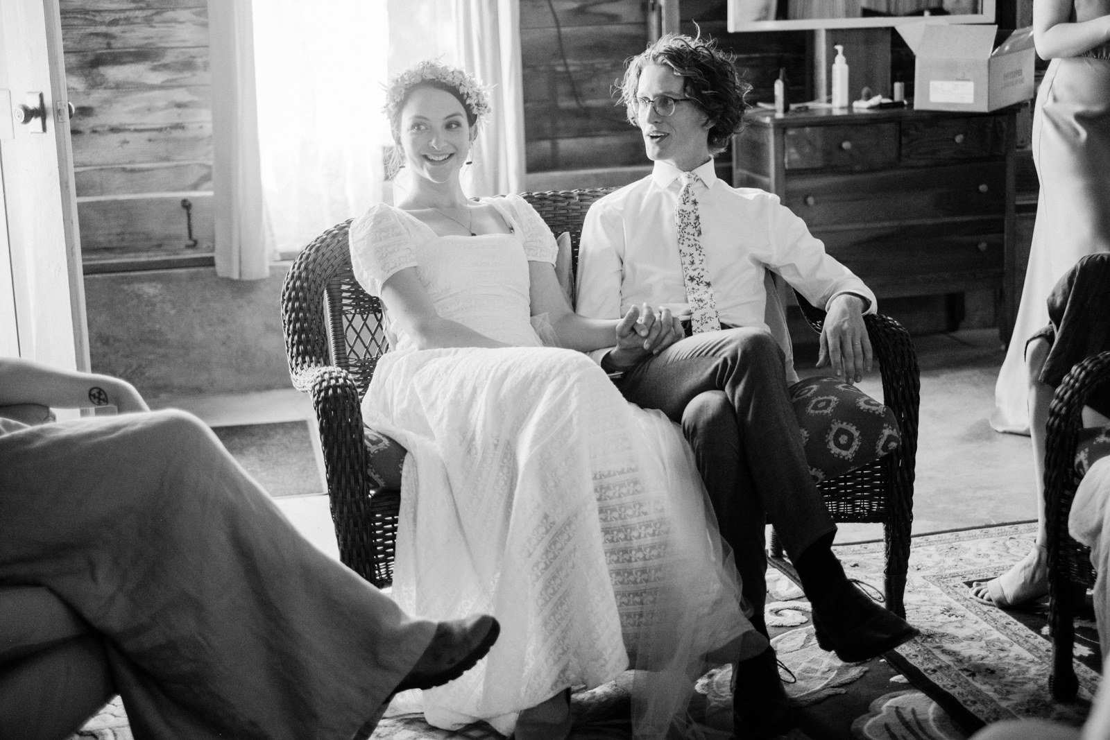  Candid moment of bride and groom sitting in wicker chair laughing with friends before wedding 