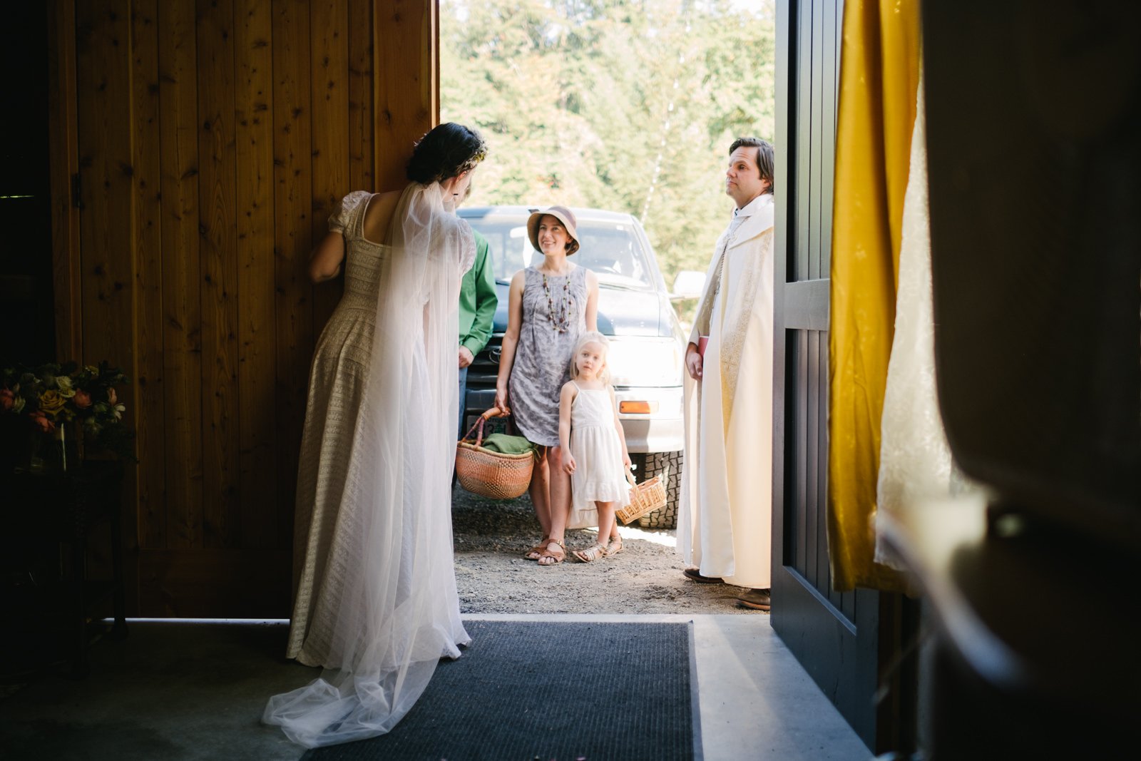  Bride peers around corner of barn waiting for efficient and mother with basket 