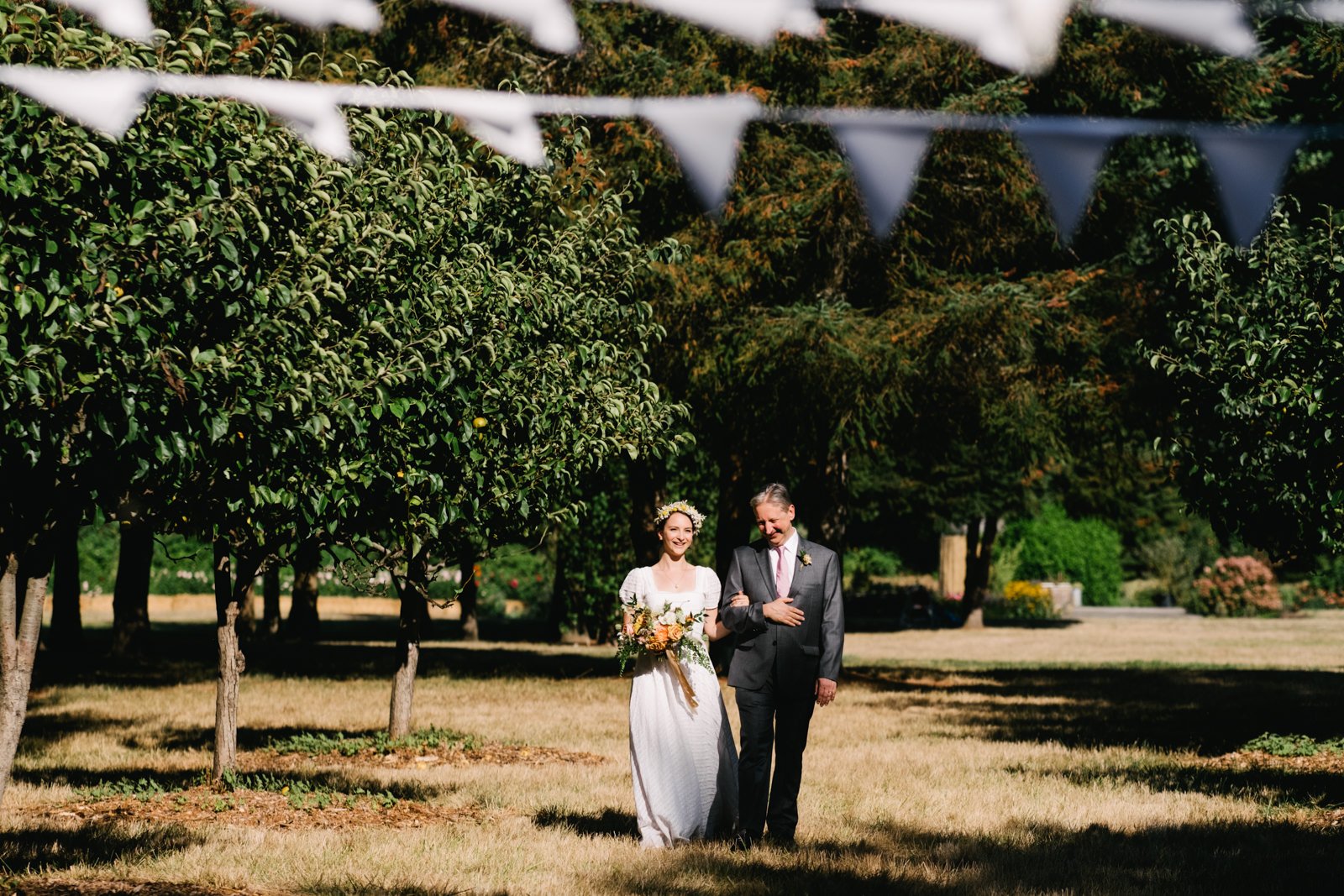  Bride walks in sunshine with father laughing in brown farm field 