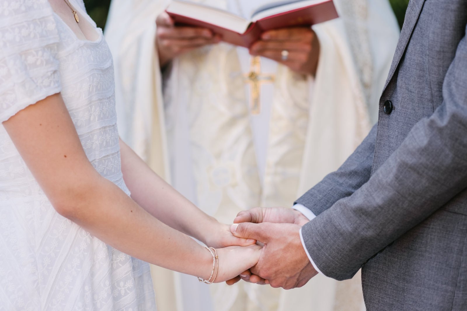  Bride and groom hold hands close up while officiant holds red bible behind them 