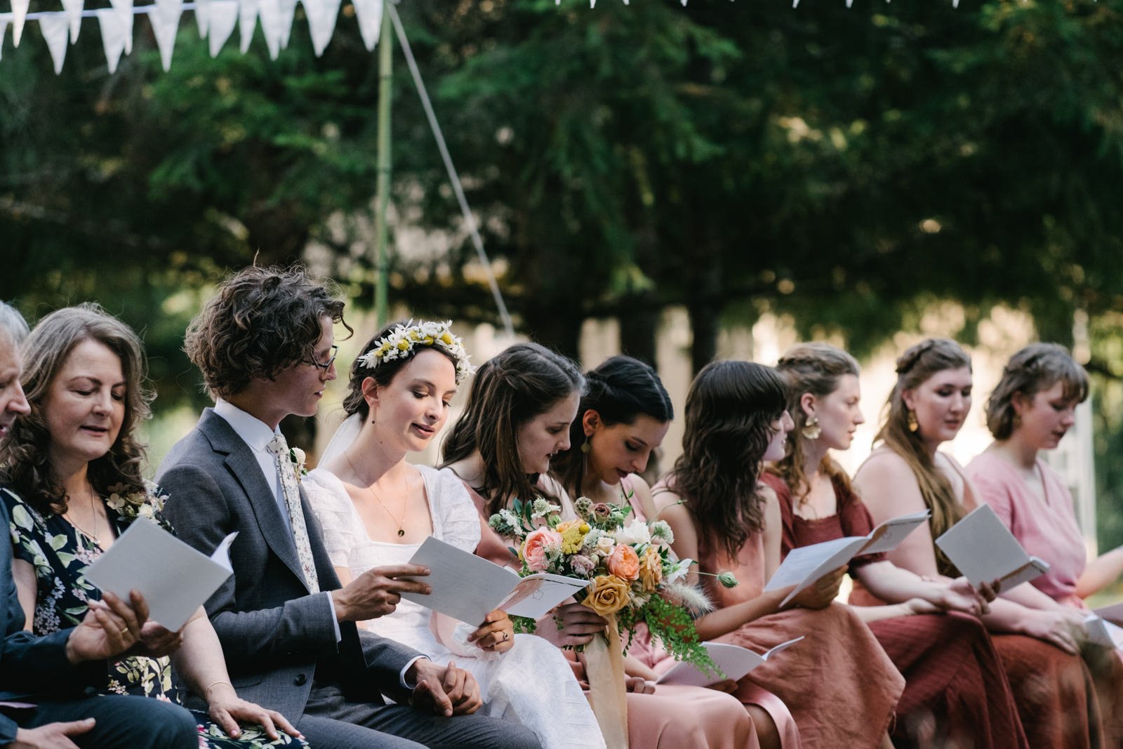  Bride and groom sing together while sun shines alone on their face while guests read hymn words 