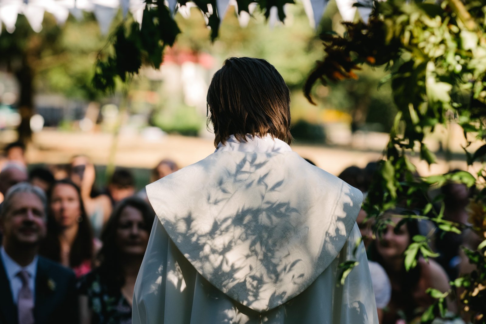  Officiant faces wedding guests while sun shines on the shade shapes of foliage and leaves pattern 