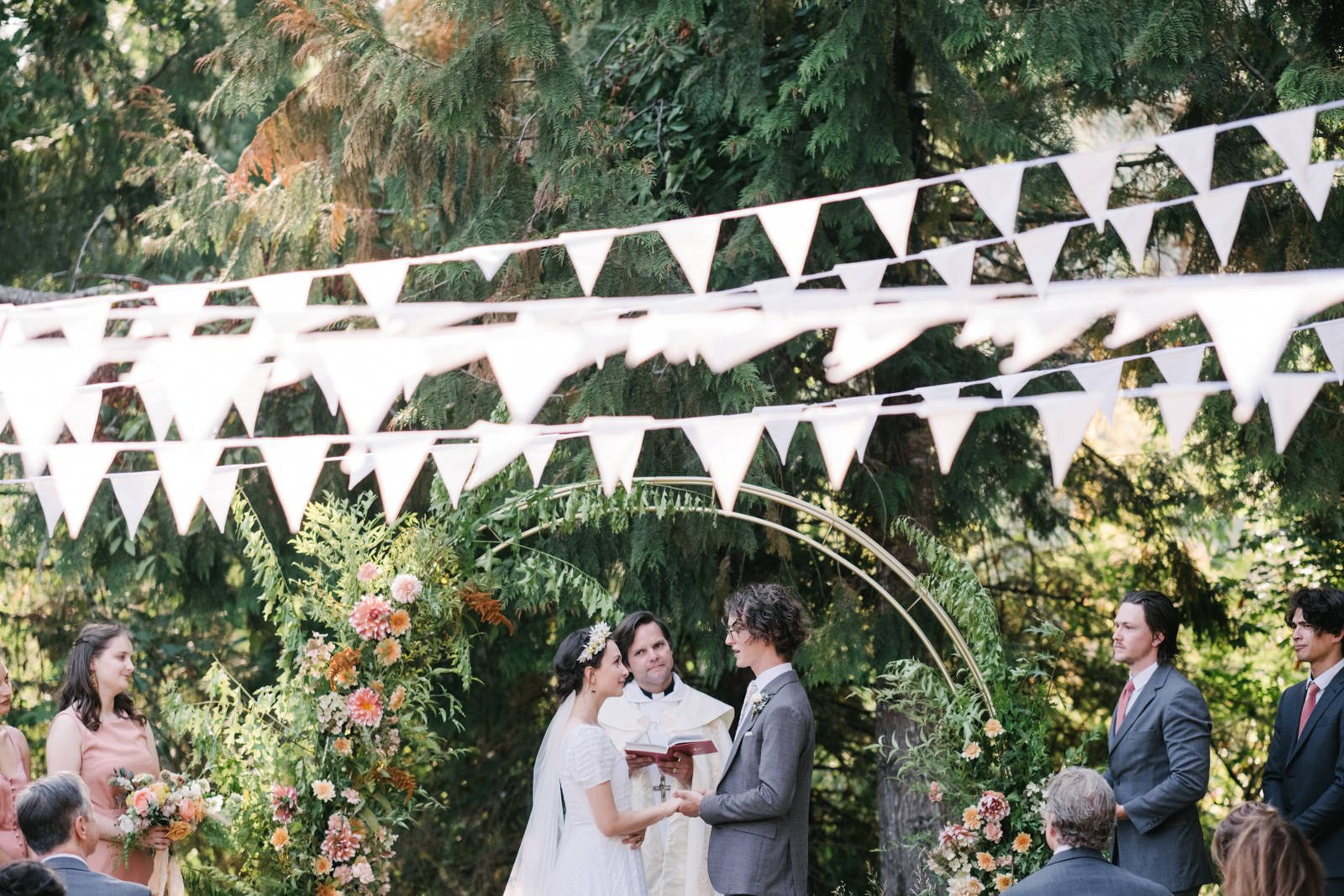  Bride and groom share vows in front of floral arch with white triangle flags above them 