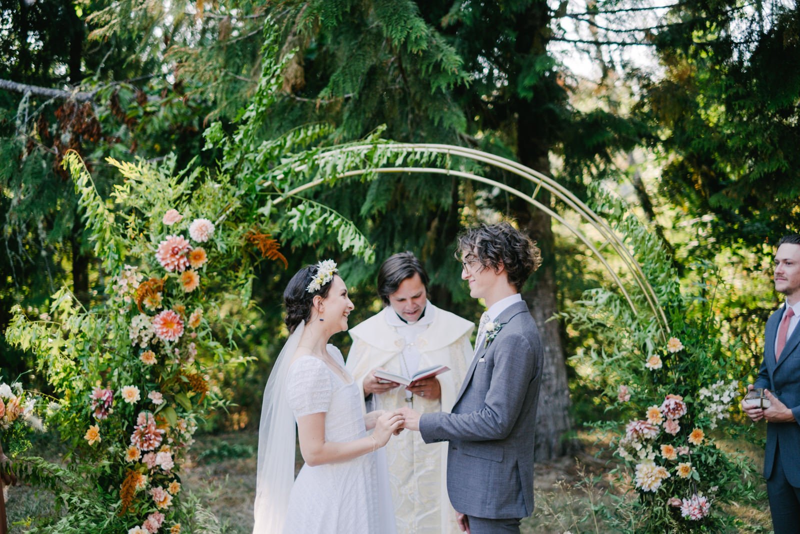  Bride places ring on groom's finger while laughing surrounded by round floral arch and fir trees 