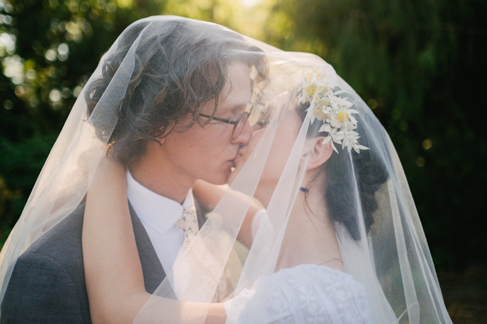  Bride and groom kiss under veil backlit by sunshine with flower crown 