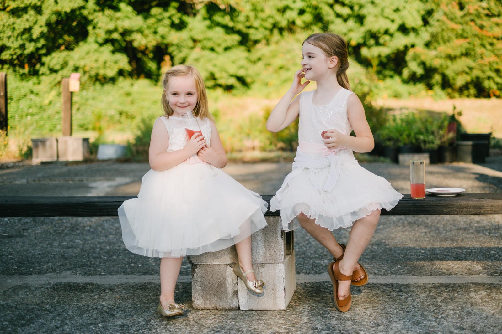  Girls in white dresses sit on bench outside during wedding reception with fruit punch 