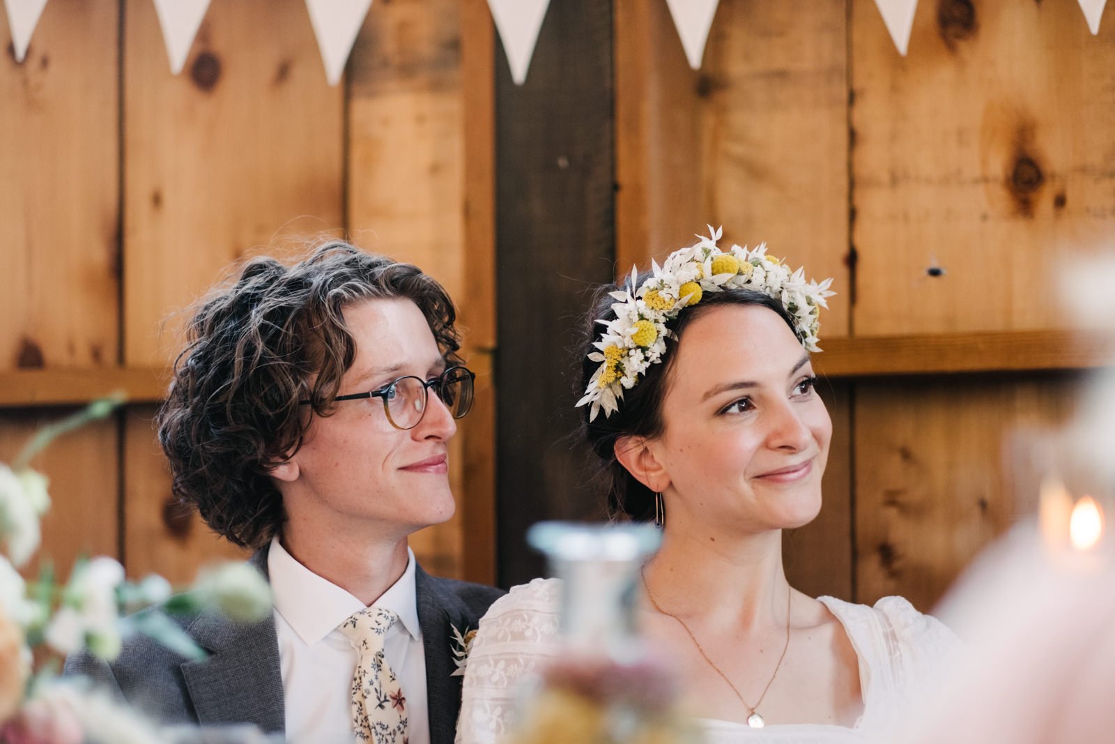  Bride and groom smile listening to toasts wearing flower crown 