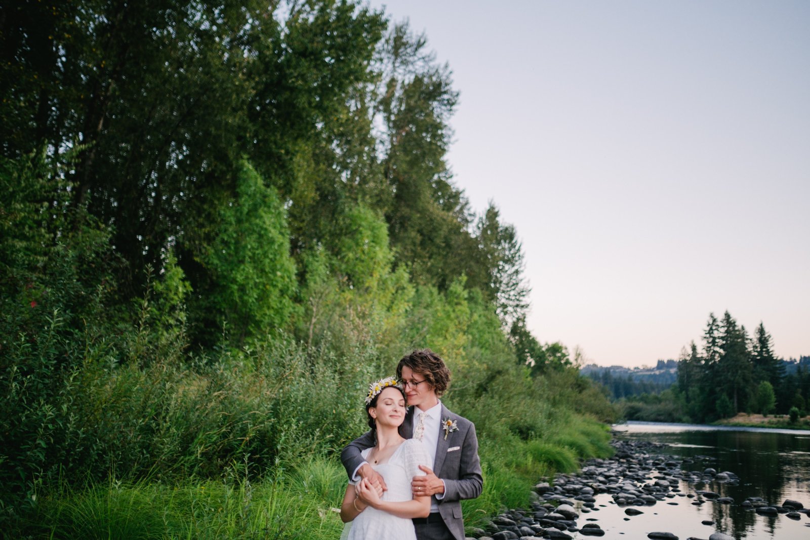  Wedding couple embrace by river in late sunset time with tall green trees 