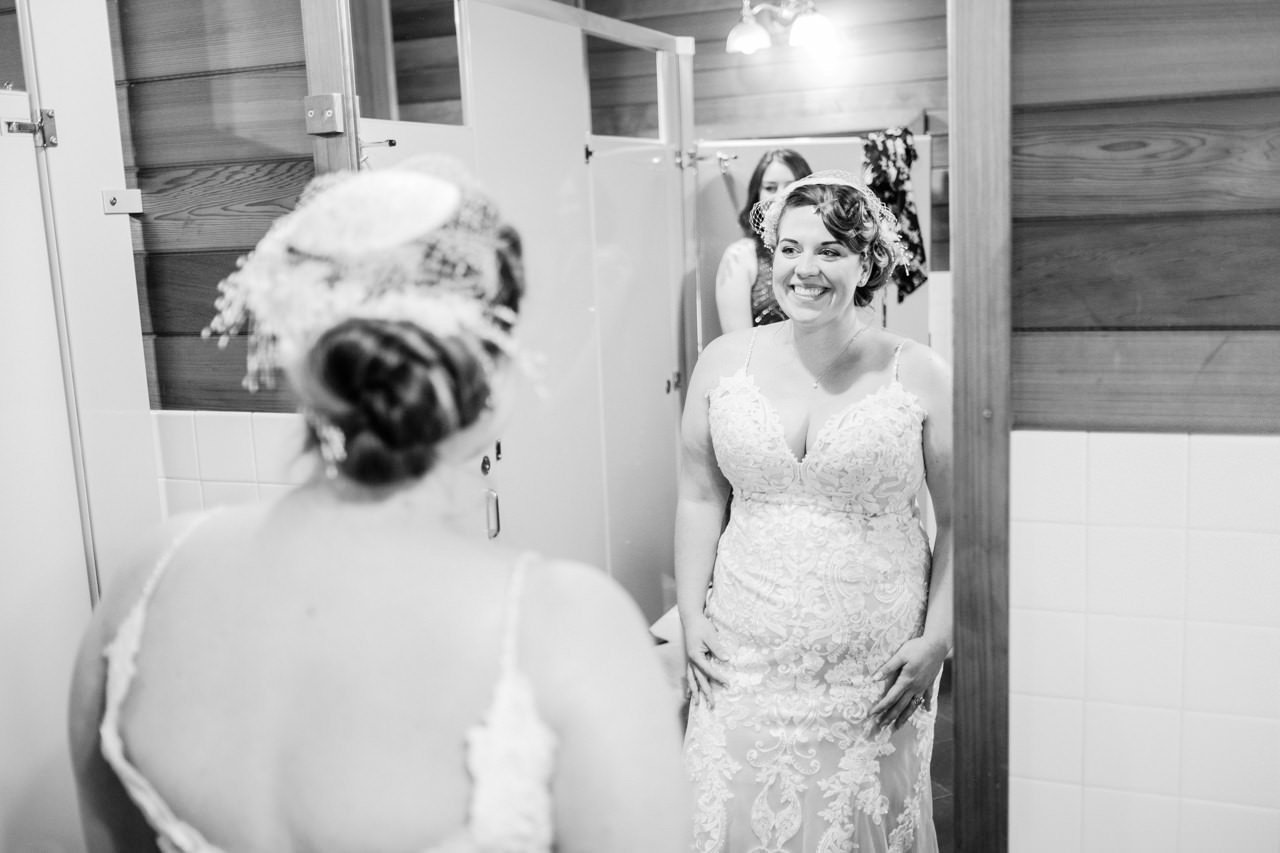  Bride with white hairpiece smiles in mirror in bathroom 