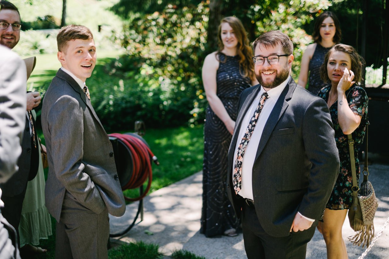  Groom in floral tie smiles at camera in candid photograph with wedding party 