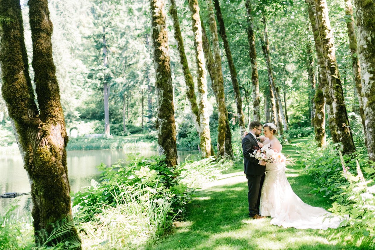  Wedding couple snuggles together by lake surrounded by grove of trees 