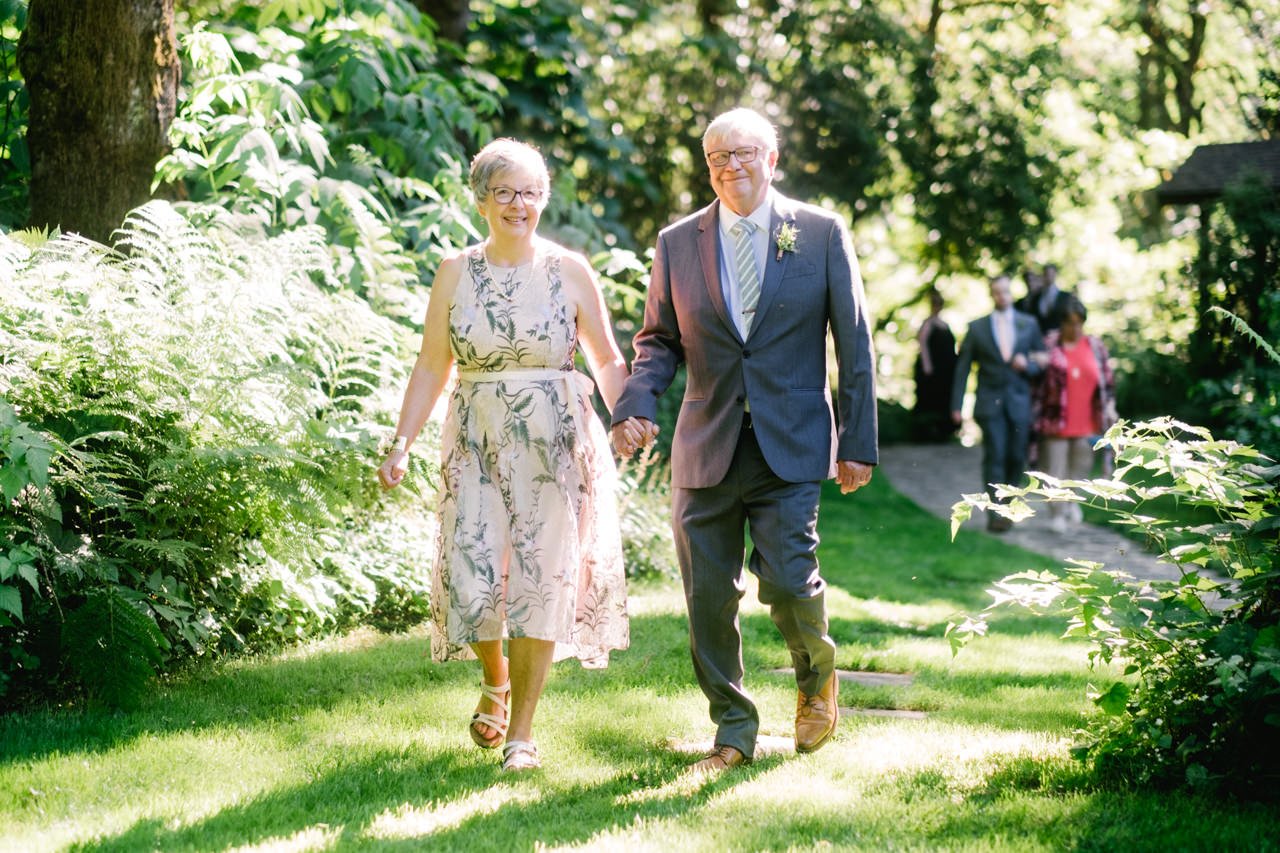  Grooms parents walk in sunlight along stone path towards ceremony 