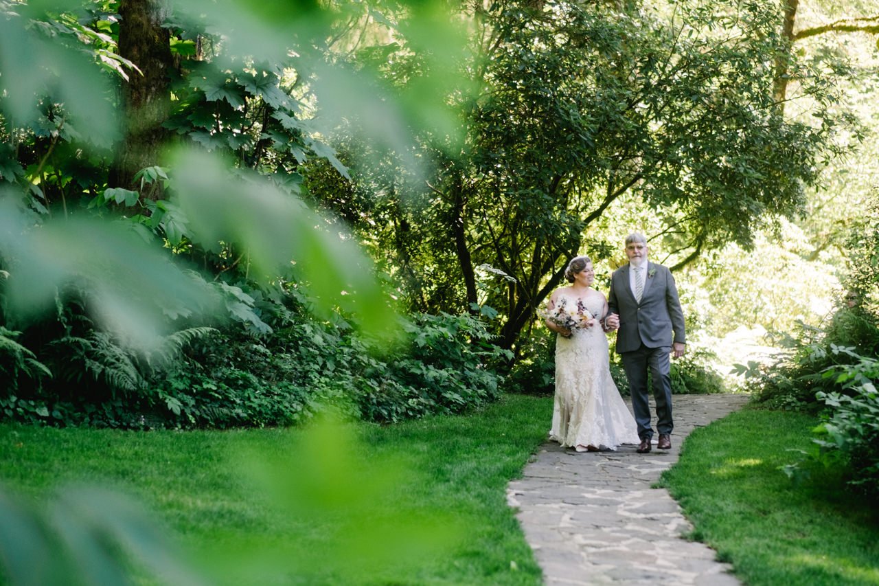  Bride walks with father in grey suit along stone path towards ceremony with foliage in foreground 