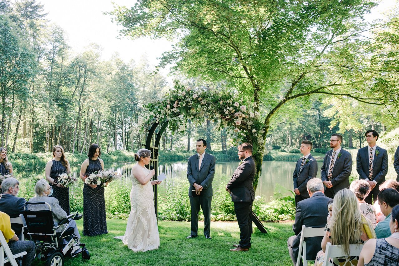  Bride shares vows in front of floral arch by lake 