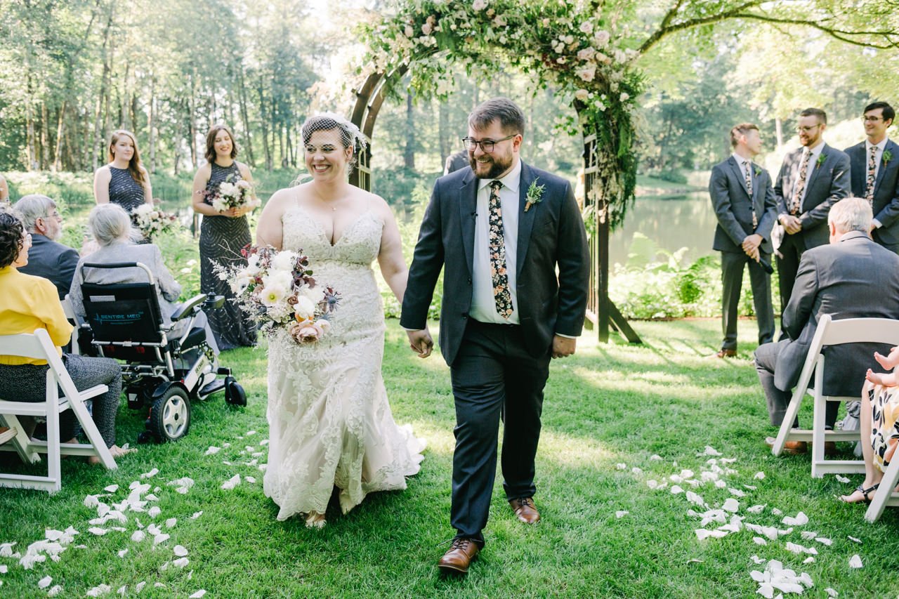  Bride and groom walk down aisle in white pedals while groom in floral tie laughs 