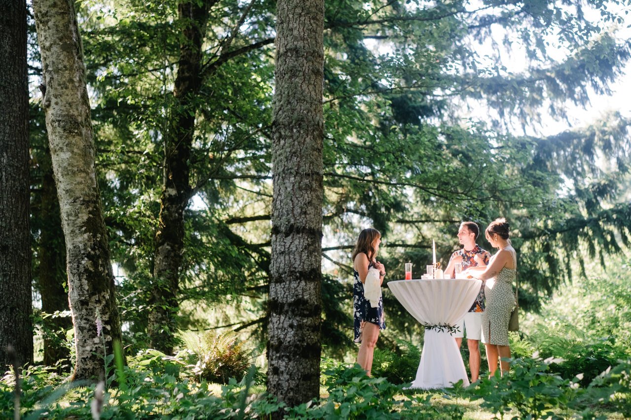  Wedding guests stand in sunlight set in forest table 