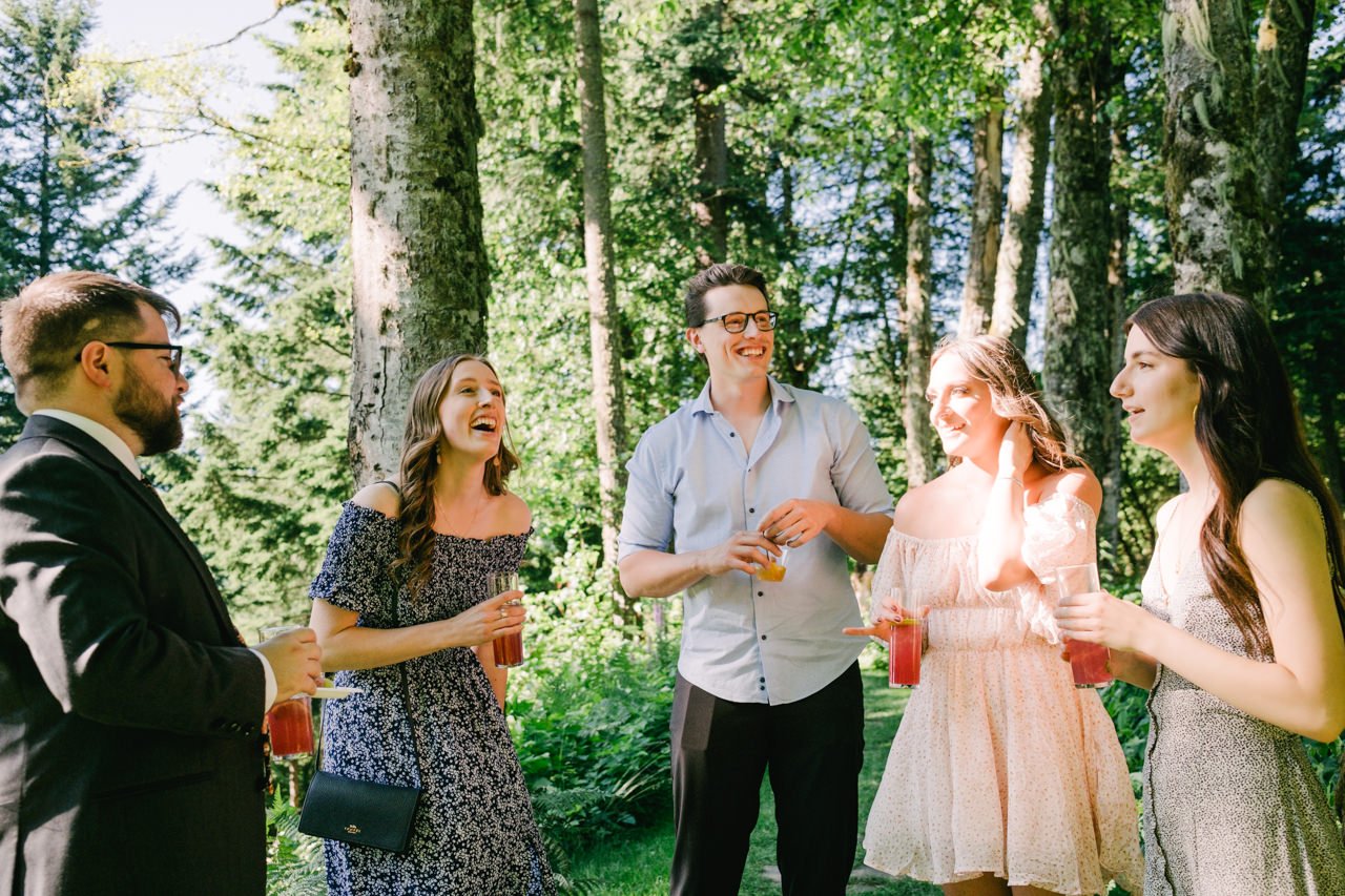  Wedding guests laughing in sunlit forest holding pink drinks during cocktail hour 